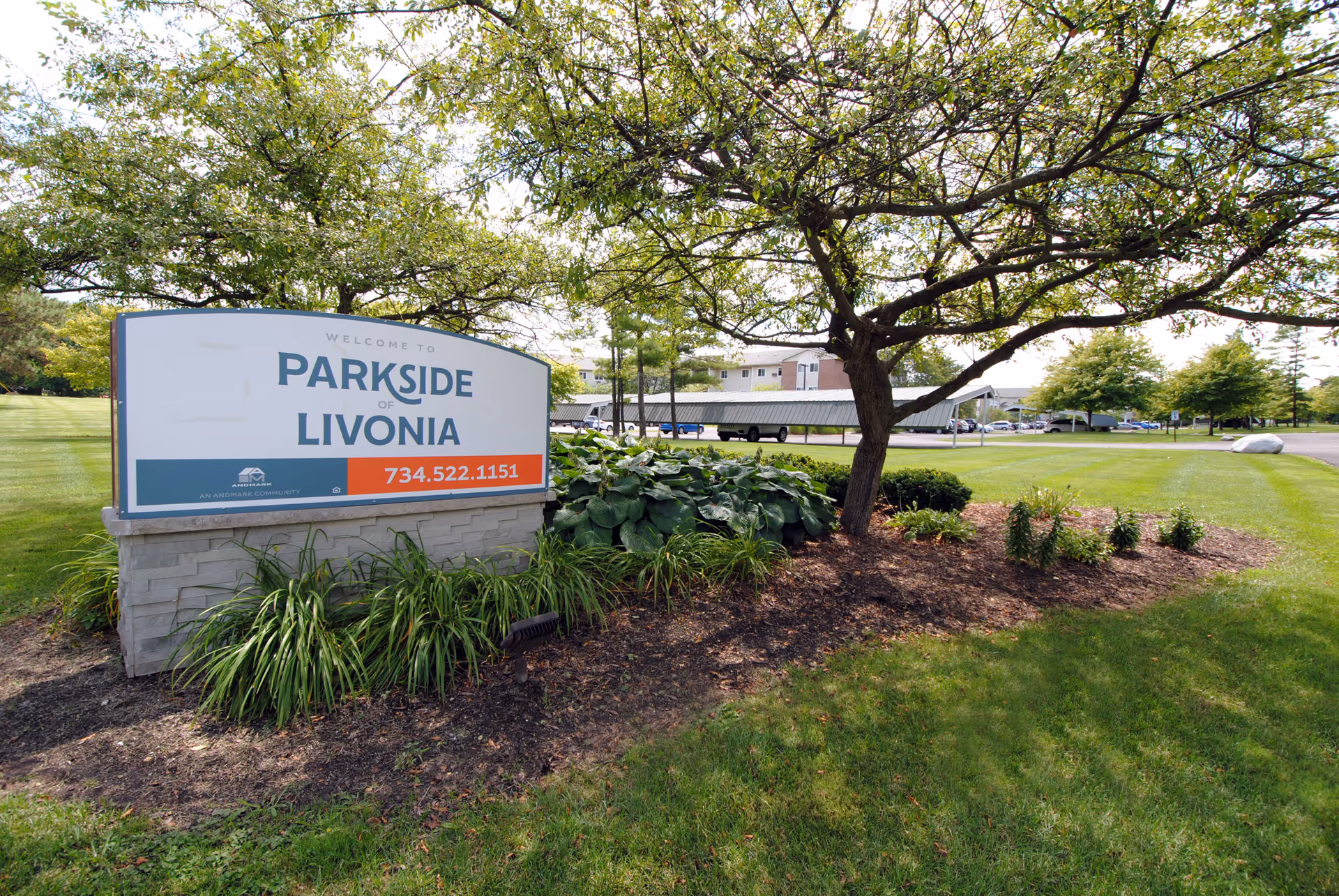 Outdoor view of the Parkside of Livonia facility sign surrounded by greenery, plants, and a tree, with a parking lot and building visible in the background.