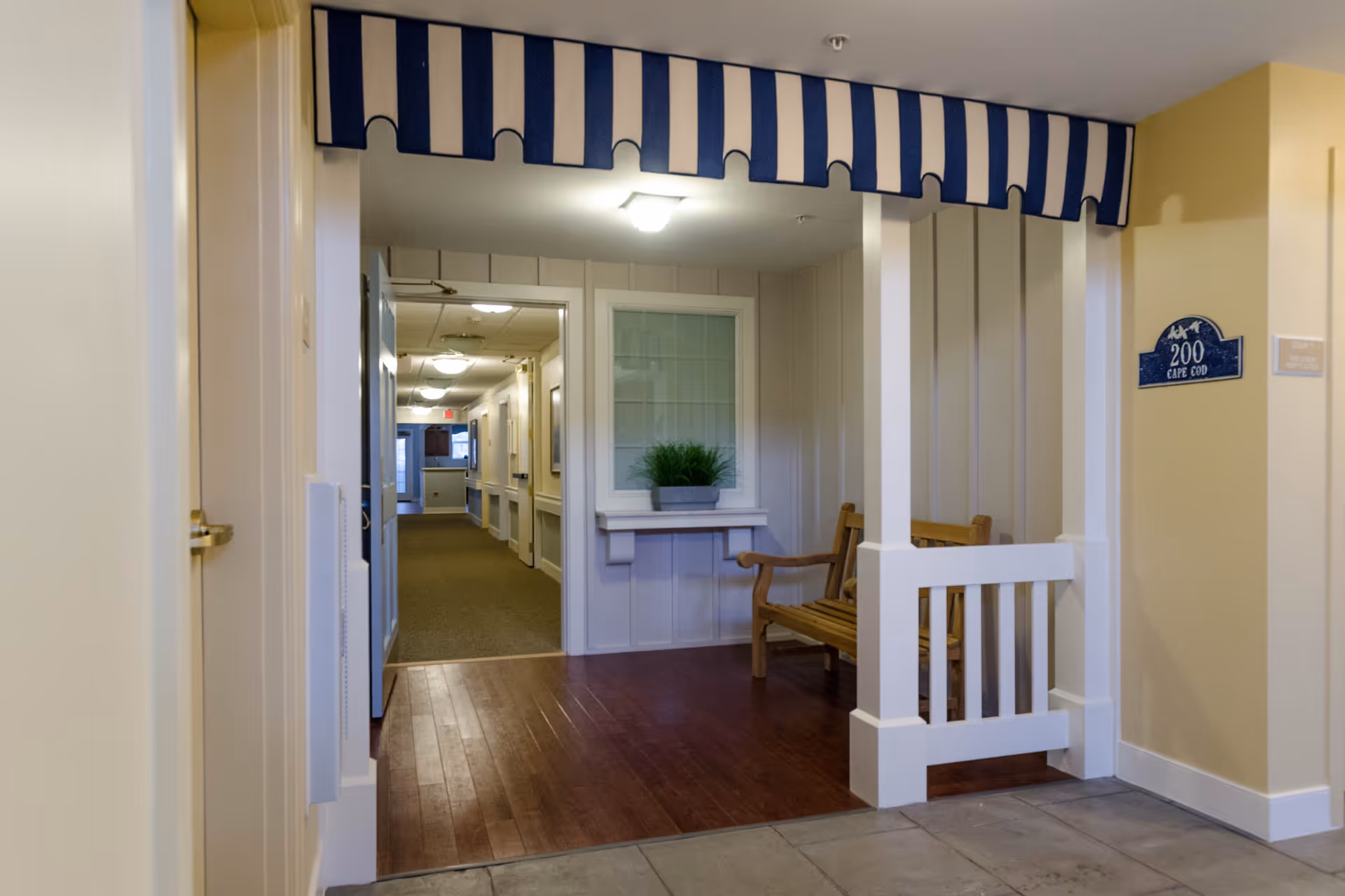 Indoor hallway area in a senior living facility with a small seating nook featuring a wooden bench, a potted plant on a shelf, and a blue and white striped canopy overhead. The hallway extends into the distance with doors and framed pictures on the walls.
