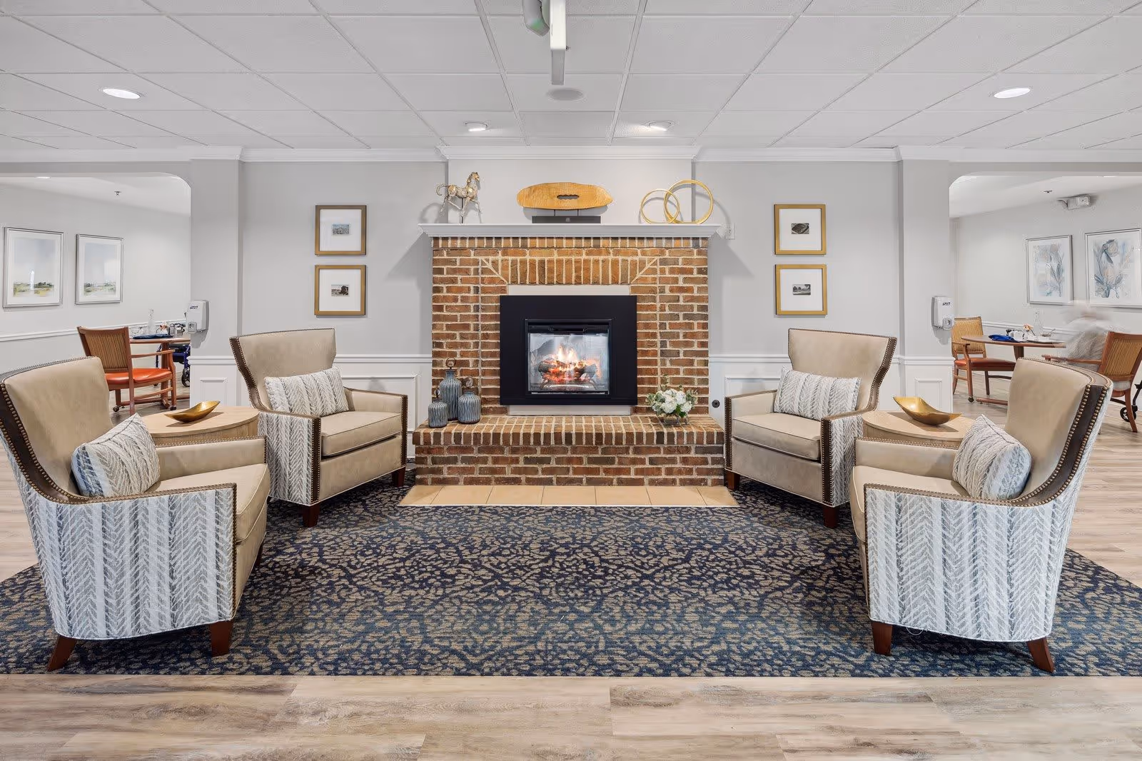 A cozy seating area with four beige armchairs arranged around a brick fireplace with a fire burning inside. The fireplace mantel is decorated with small sculptures and framed pictures hang on the wall. The floor has a patterned blue rug and light wood flooring. In the background, there are dining tables and chairs in a well-lit room.