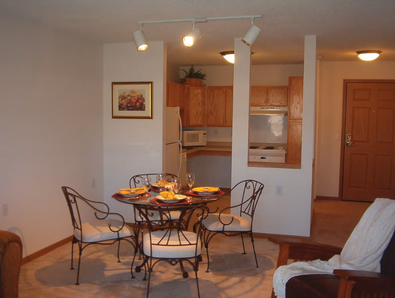 Interior view of a living and dining area with a round glass dining table set for four with yellow napkins and glassware. The dining area is adjacent to a kitchen with wooden cabinets, a white refrigerator, microwave, stove, and dishwasher. There is a brown armchair with a white throw blanket on the right side and a wooden front door in the background.