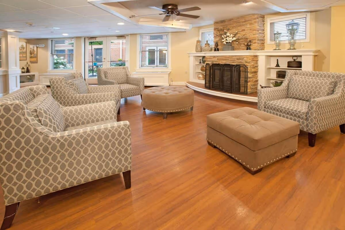 A cozy common lounge with patterned armchairs, tufted ottomans, hardwood floors, and a stone fireplace.
