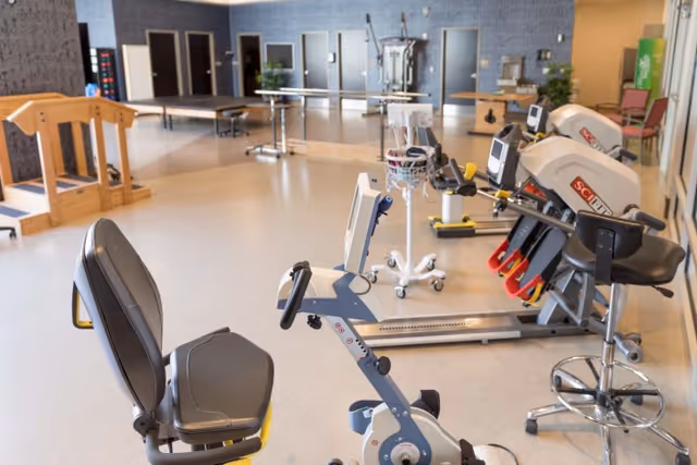 A spacious rehabilitation or physical therapy room with exercise equipment including stationary bikes and other fitness machines. The room has a light-colored floor, several doors along the back wall, and a few tables and chairs. The environment is clean and well-lit.