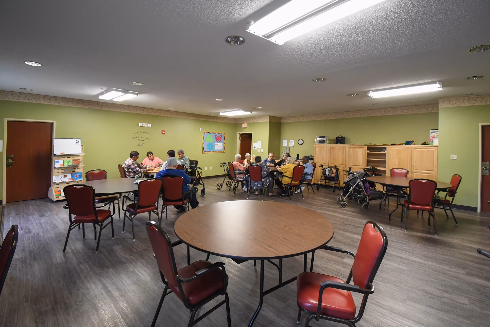 A group of elderly people sitting around tables in a spacious room with green walls and wooden flooring, some using walkers. The room has multiple round tables with red cushioned chairs and fluorescent ceiling lights.