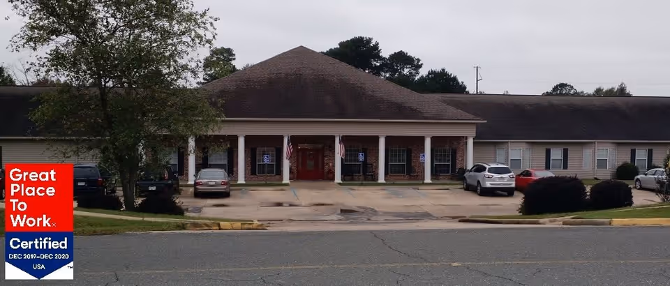 Front exterior view of The Columns of Germantown Assisted Living Community building with a covered entrance supported by white columns, several parked cars, and two American flags near the entrance.
