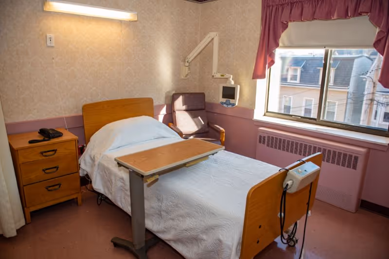 A nursing home bedroom with a single bed covered in a white quilt, a wooden bedside table with a telephone, a reclining chair, a wall-mounted medical monitor, and a window with pink curtains letting in natural light.
