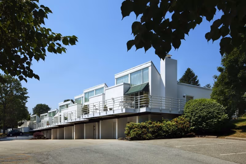 Modern white multi-unit building with elevated balconies and carports surrounded by trees under a clear blue sky.