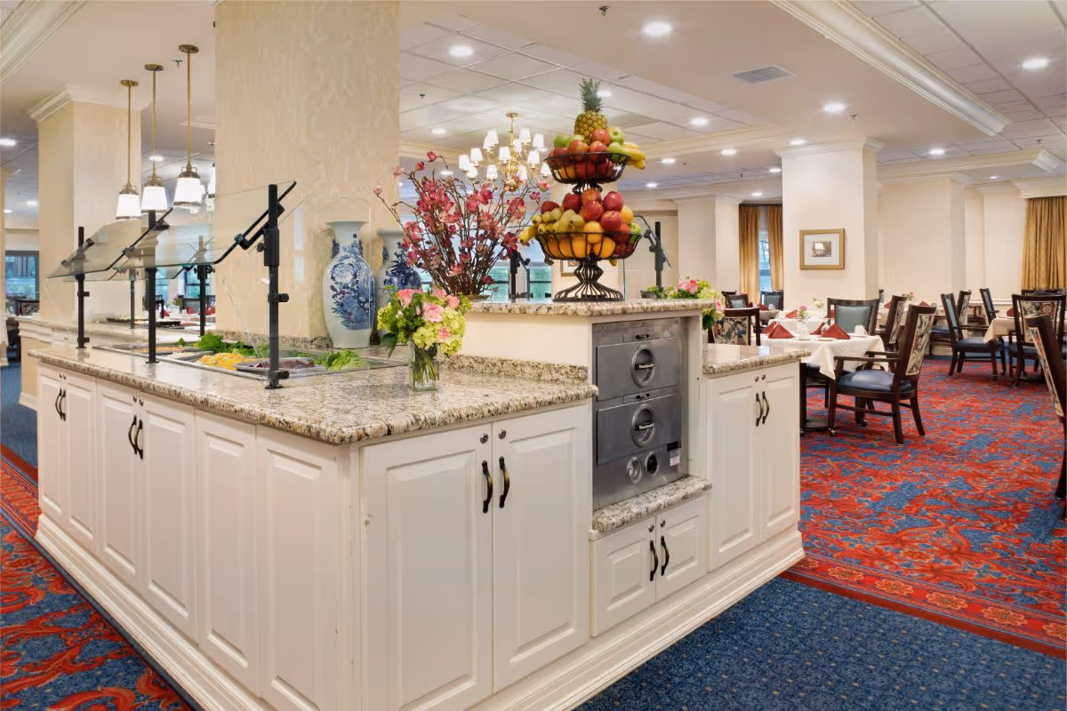 A dining area in a senior living facility featuring a buffet counter with a granite countertop, glass sneeze guards, and trays of food. The counter is decorated with a tiered fruit basket and floral arrangements. In the background, there are tables set with white tablecloths, red napkins, and chairs, all on a red and blue patterned carpet under warm lighting.
