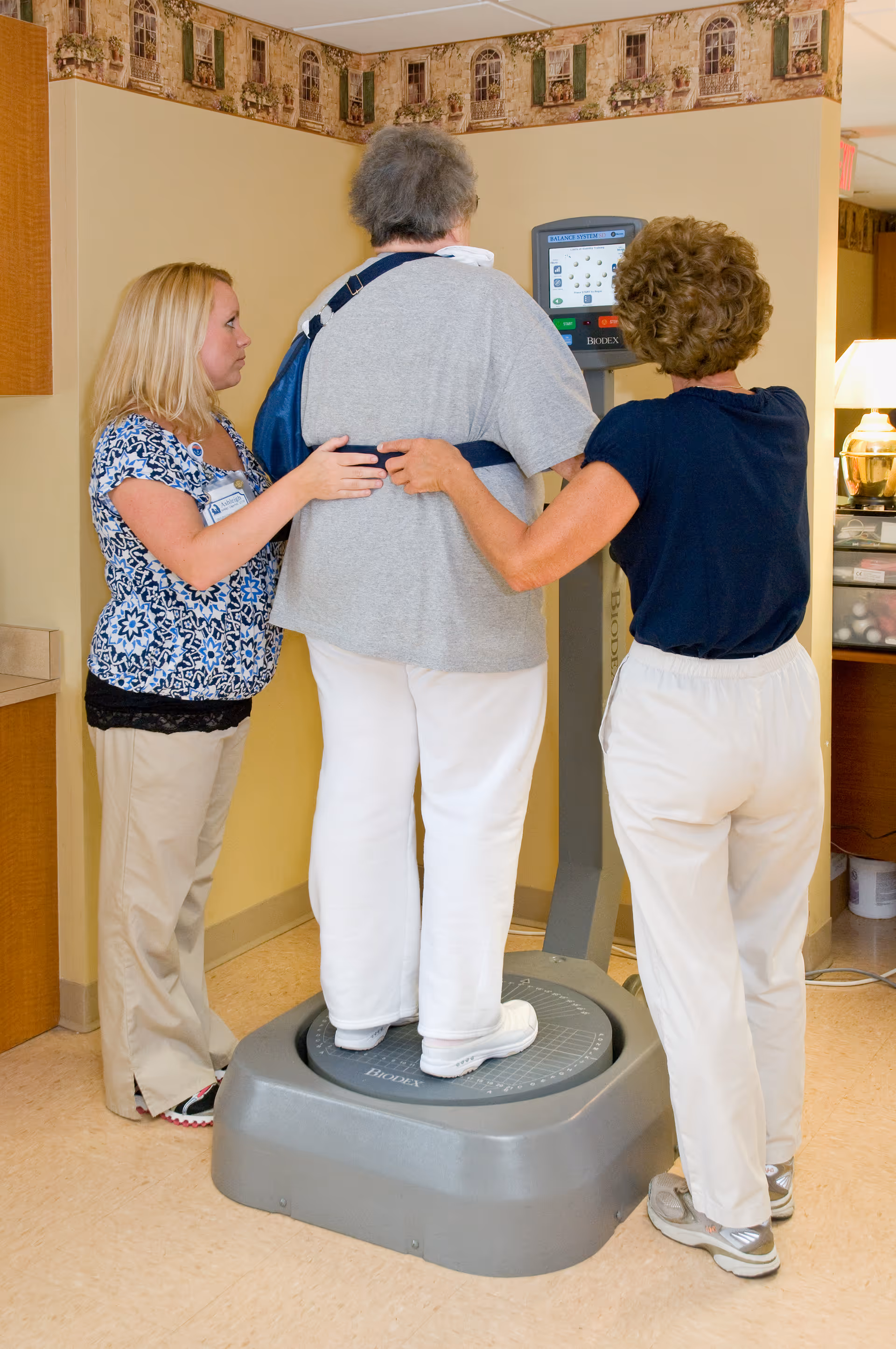 An elderly person wearing a gray shirt and white pants stands on a balance testing machine while being supported by two caregivers, one on each side, in a room with beige walls and a decorative border near the ceiling.