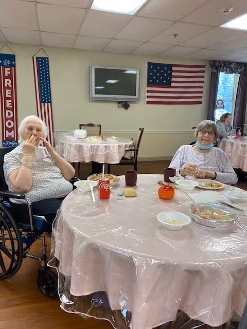 Two elderly women sitting at a round table covered with a pink plastic tablecloth in a dining area. One woman is in a wheelchair eating, and the other is smiling with a mask under her chin. The table has plates of food, a can of Coca-Cola, cups, and a small pumpkin decoration. In the background, there are other tables with similar settings, an American flag on the wall, a flat-screen TV, and a vertical banner with the word 'FREEDOM'.