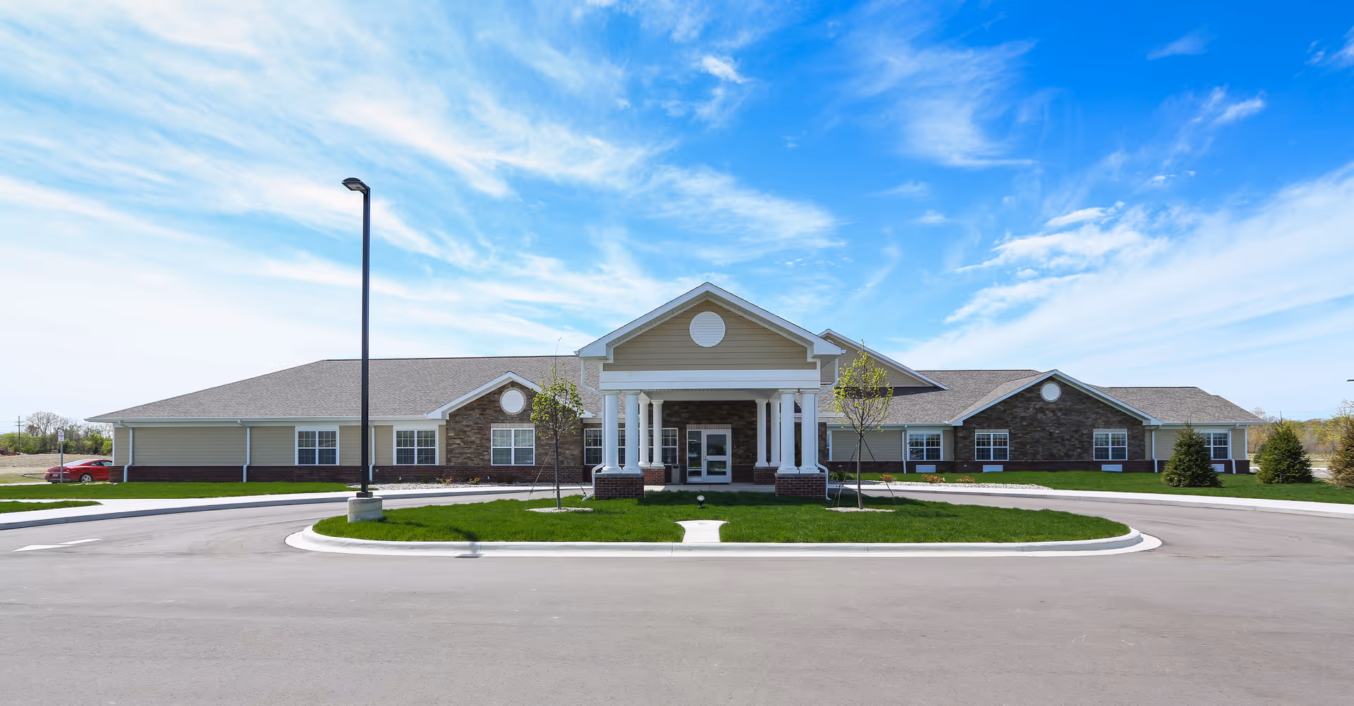 Front exterior of a single-story senior living building with a columned entrance, circular driveway, and green lawn under a blue sky.