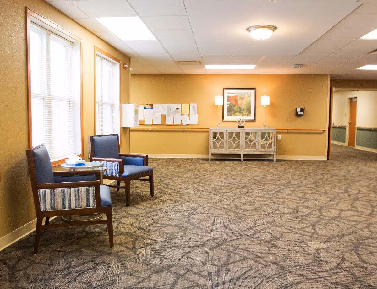 A quiet hallway area in a senior living facility with two blue cushioned chairs and a small round table near two large windows with blinds. The walls are painted beige and decorated with a framed picture, two lamps on a decorative cabinet, and a bulletin board with papers. The floor is carpeted with a patterned design.