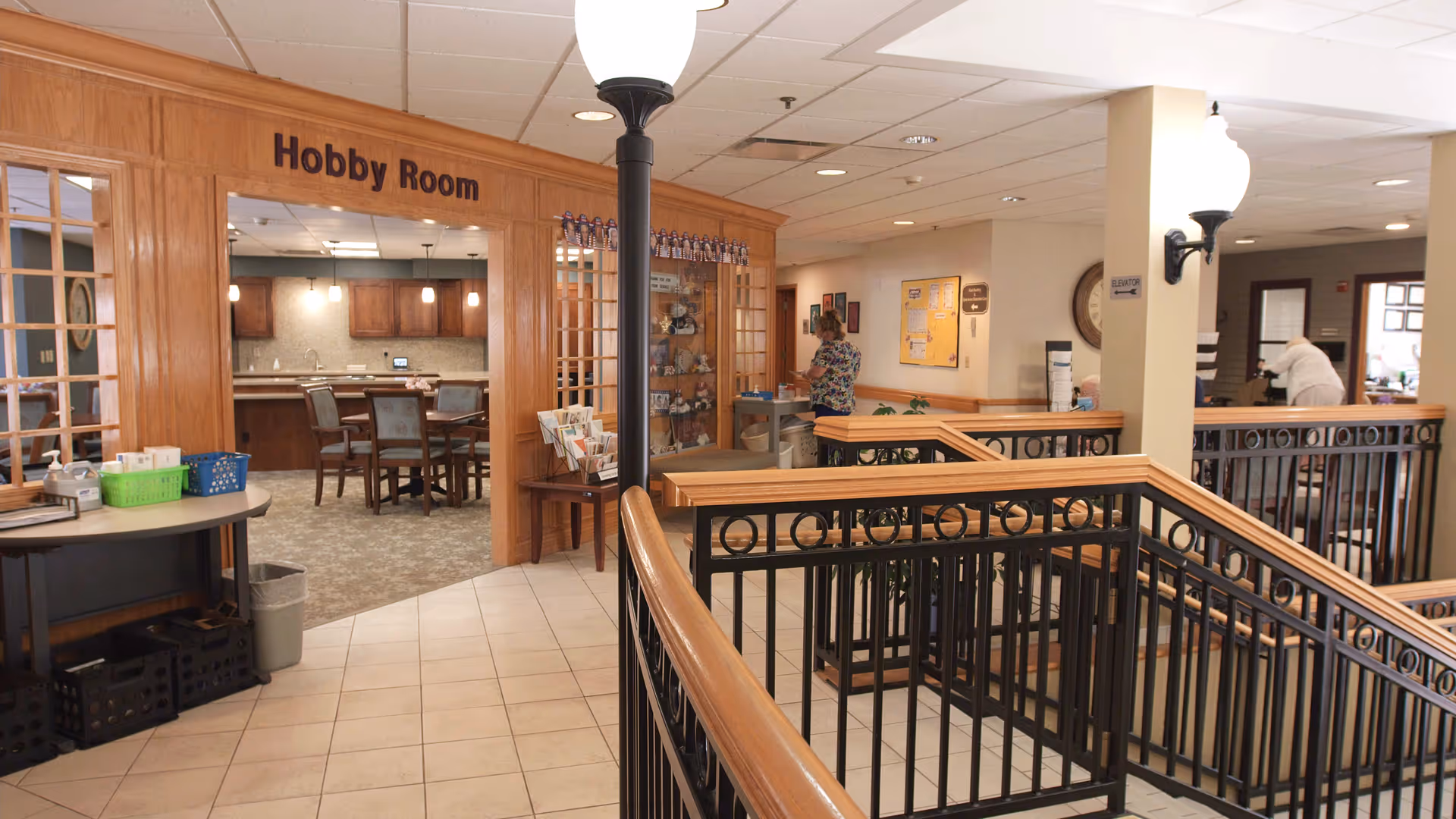 Interior view of a senior living facility showing a hallway with a railing and a staircase. To the left is an entrance labeled 'Hobby Room' with wooden framing and glass panels. Inside the hobby room, there are tables and chairs with a kitchen area in the background. Two people are visible in the hallway and another room further back. The area is well-lit with ceiling lights and wall-mounted lamps.