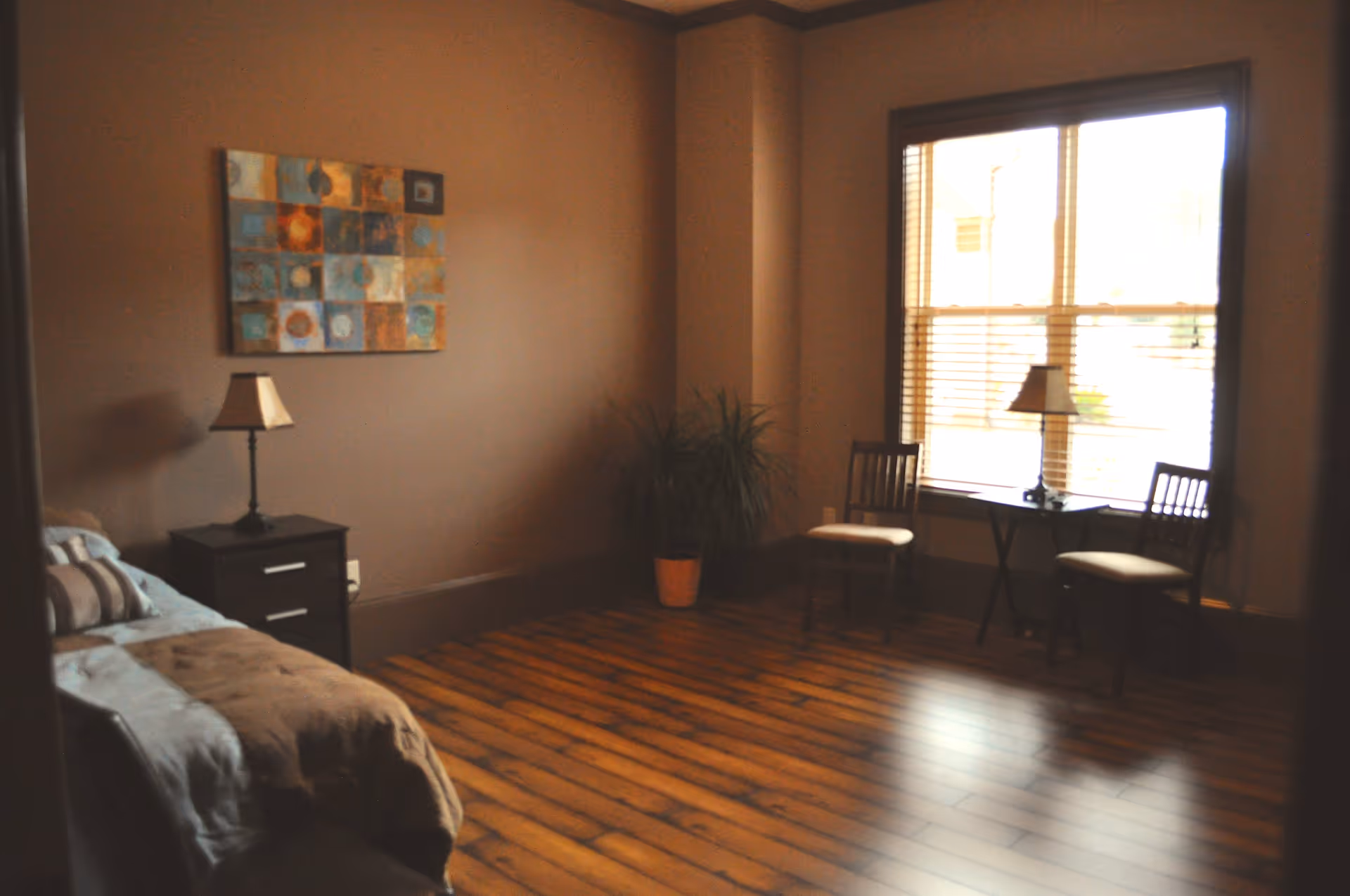 A cozy room with wooden flooring, a bed with pillows and a blanket on the left, a nightstand with a lamp, a potted plant in the corner, and two chairs with a small table and lamp near a large window with blinds.