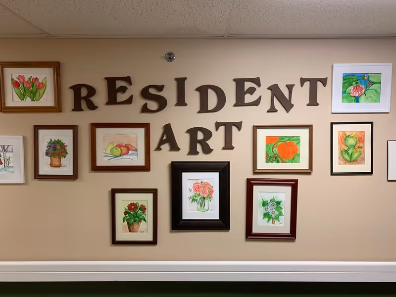 A beige wall decorated with framed watercolor paintings of flowers, fruits, and plants. Above the paintings, large brown letters spell out 'RESIDENT ART'.