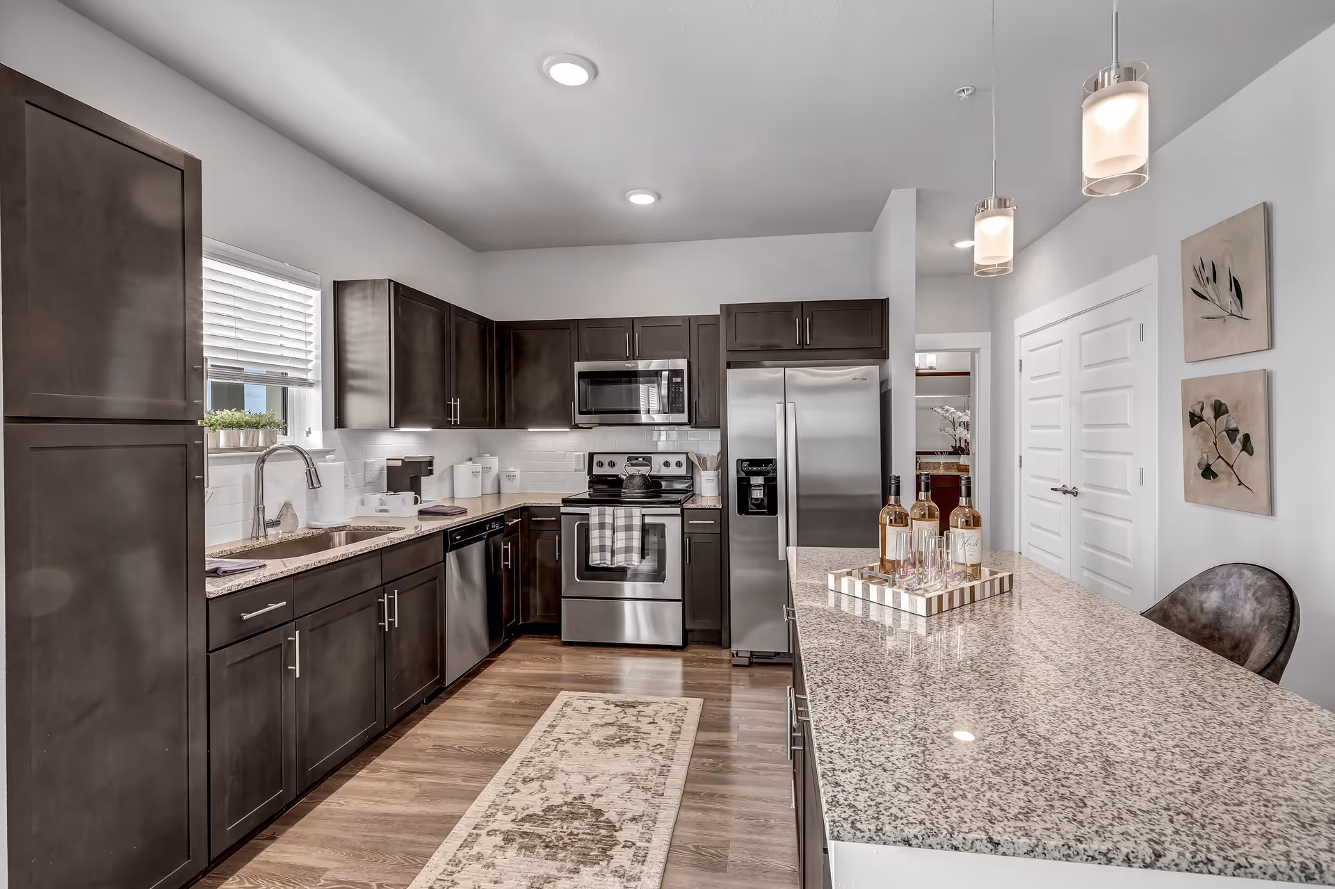 Modern kitchen with dark wood cabinets, stainless steel appliances, and a granite island topped with bottles and pendant lights.