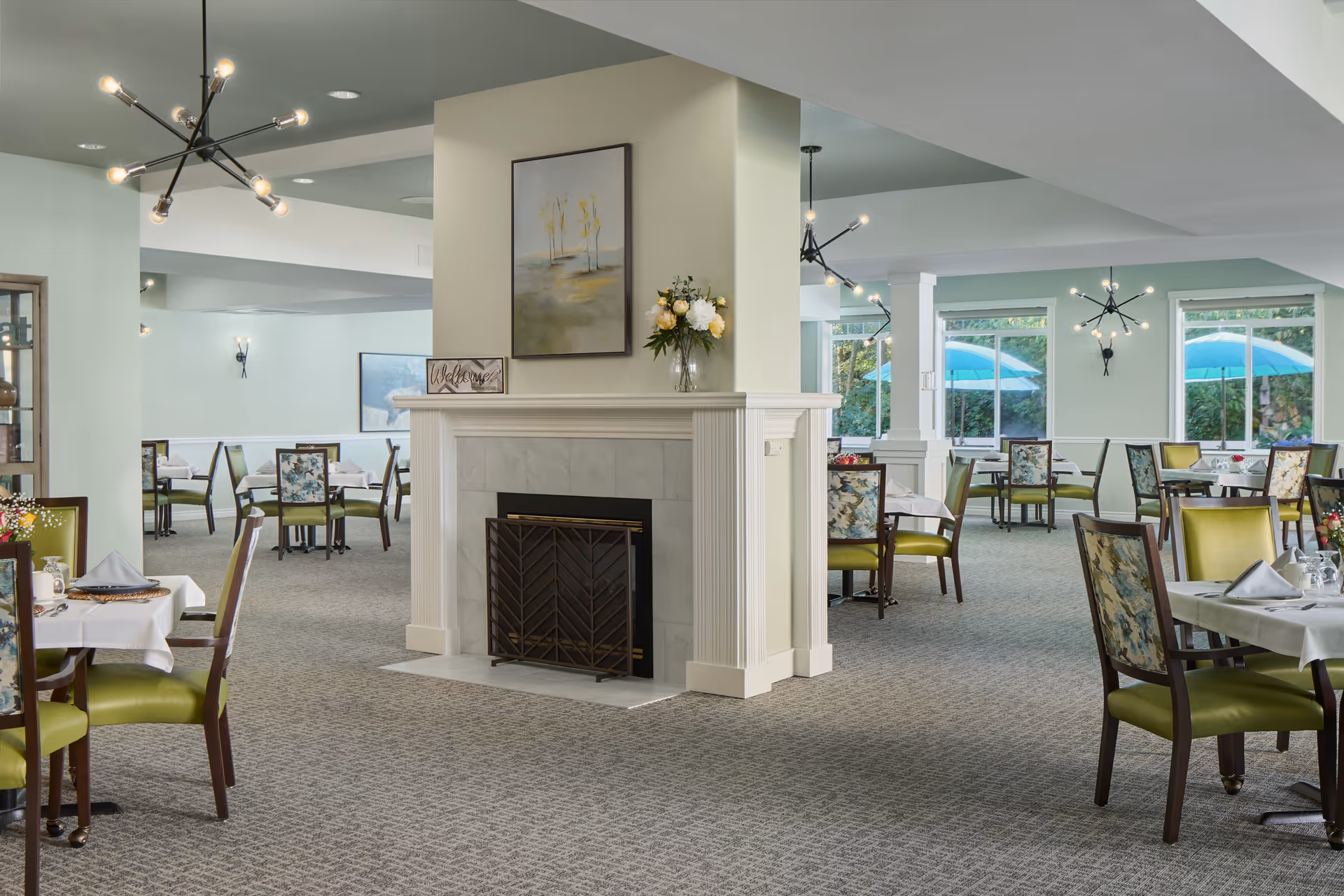 Dining room with multiple tables and green-upholstered chairs arranged around a central fireplace and large windows.
