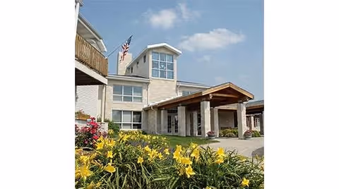 Front entrance of a senior living building with a covered porte-cochère, landscaped yellow flowers, and an American flag.