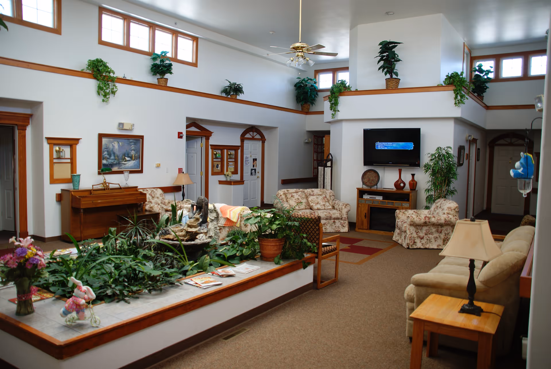 A spacious living room area in a senior living facility with multiple floral-patterned sofas and armchairs arranged around a central indoor garden with various green plants. The room features high ceilings with several windows near the top, a ceiling fan with lights, a wall-mounted flat-screen TV, wooden furniture including a piano, side tables with lamps, and decorative plants placed on ledges and corners.