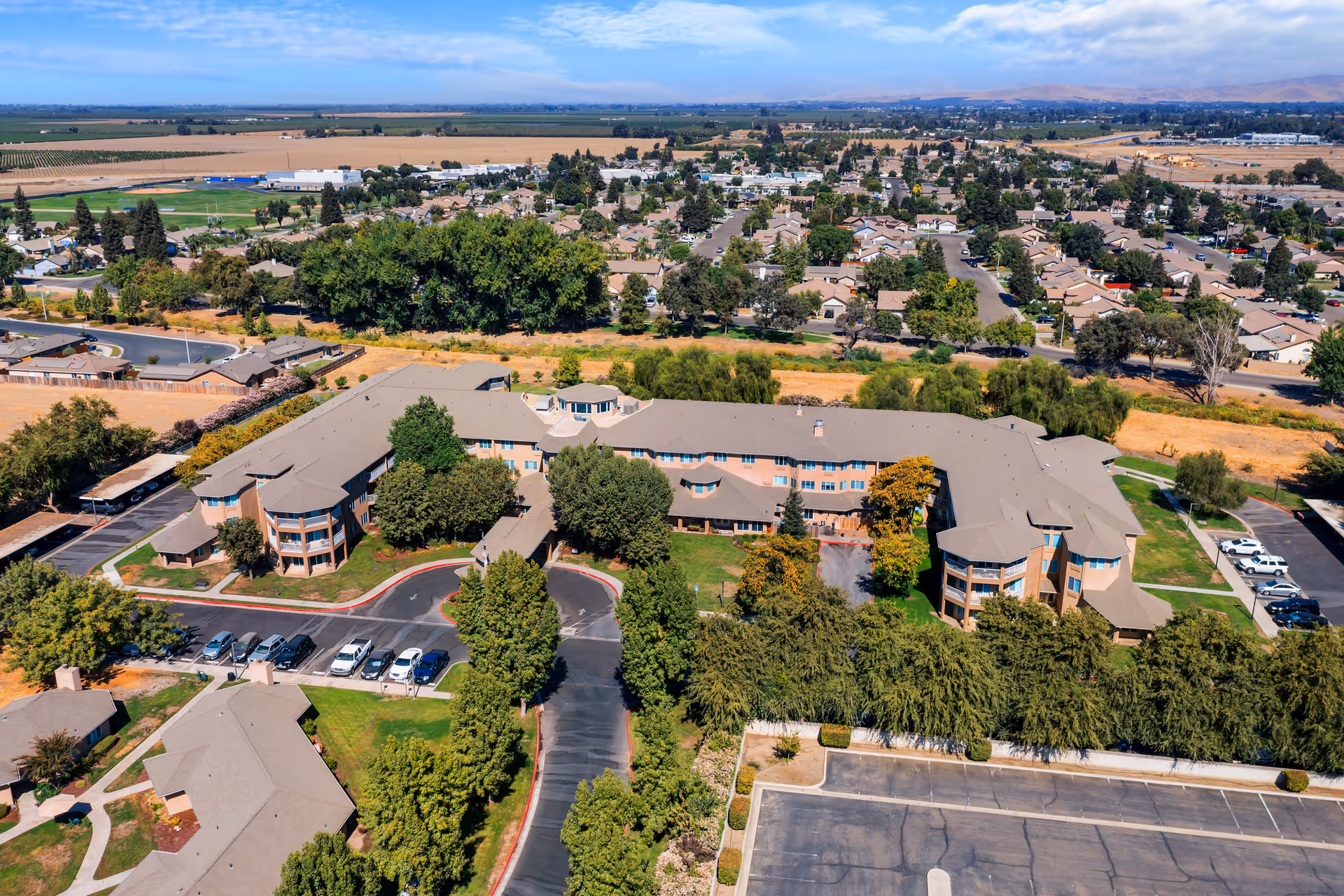 Aerial view of Sierra Hills - A Provincial Senior Living Community, showing a large, multi-wing building surrounded by trees, parking lots with cars, and nearby residential neighborhoods under a clear blue sky.