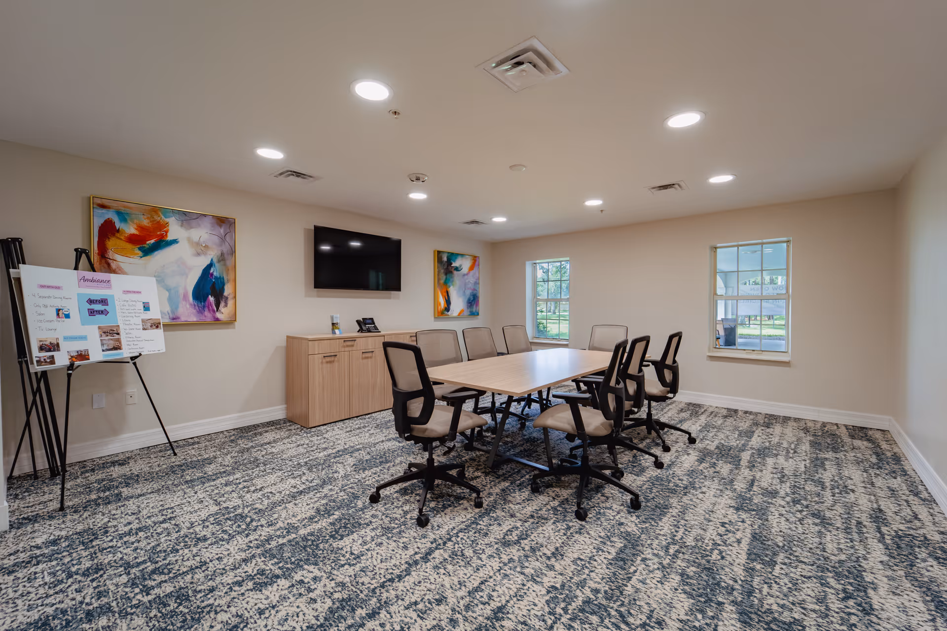 A modern conference room with a rectangular table surrounded by eight office chairs on wheels. The room has two windows letting in natural light, two colorful abstract paintings on the walls, a flat-screen TV mounted above a wooden cabinet, and a presentation board on an easel. The floor is carpeted with a patterned design and the ceiling has recessed lighting.