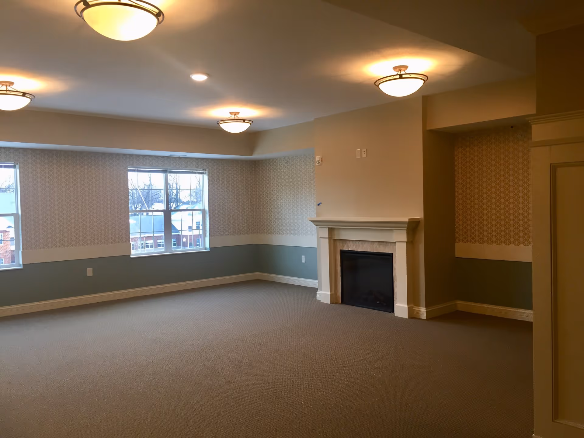 Empty carpeted common room with a fireplace, multiple ceiling lights, patterned walls and two windows.