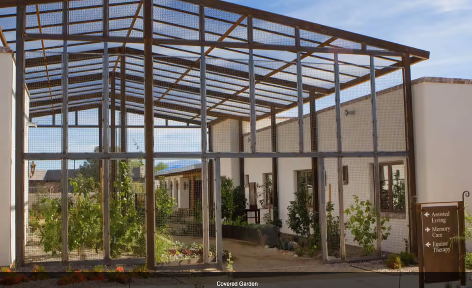 A covered garden area enclosed with a metal frame and mesh, featuring plants and greenery inside. The garden is adjacent to a white building with windows and doors. A wooden sign near the entrance points to Assisted Living, Memory Care, and Equine Therapy.