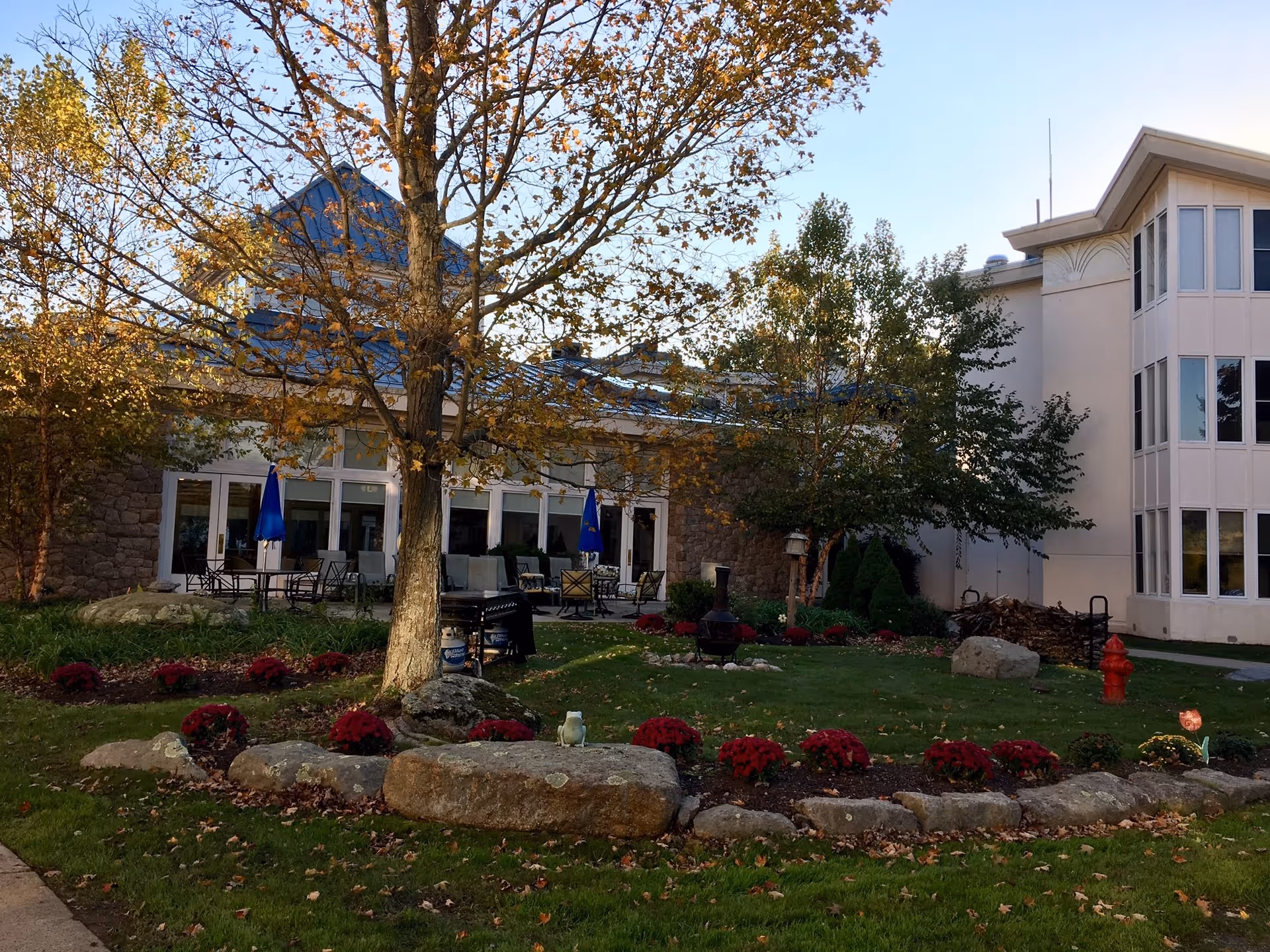 Exterior front of a retirement residence showing a landscaped lawn with trees, flower beds, rock border and a patio with tables and umbrellas.
