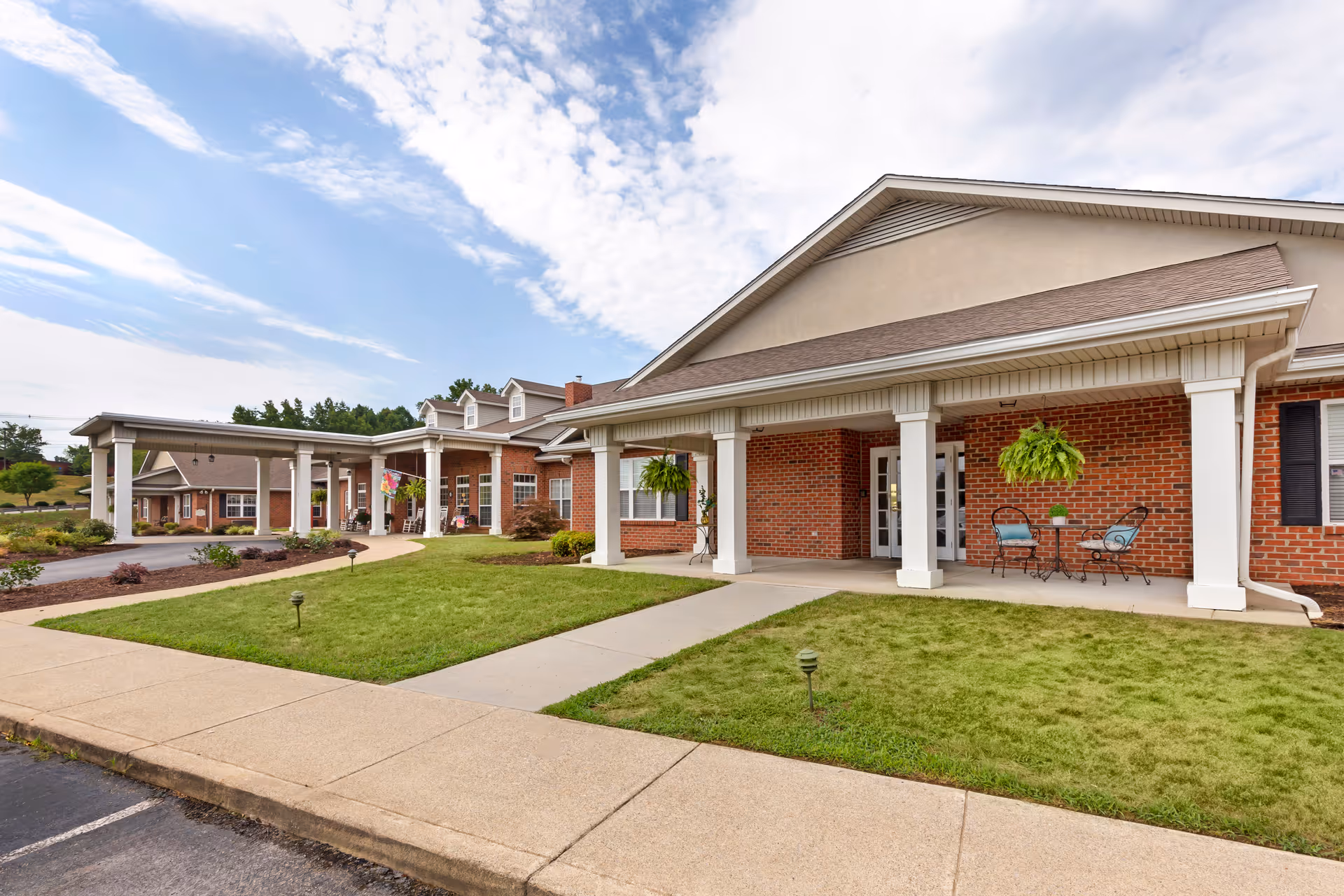 Exterior view of a senior living facility building with a covered entrance supported by white columns, red brick walls, and a well-maintained lawn with hanging plants and outdoor seating.