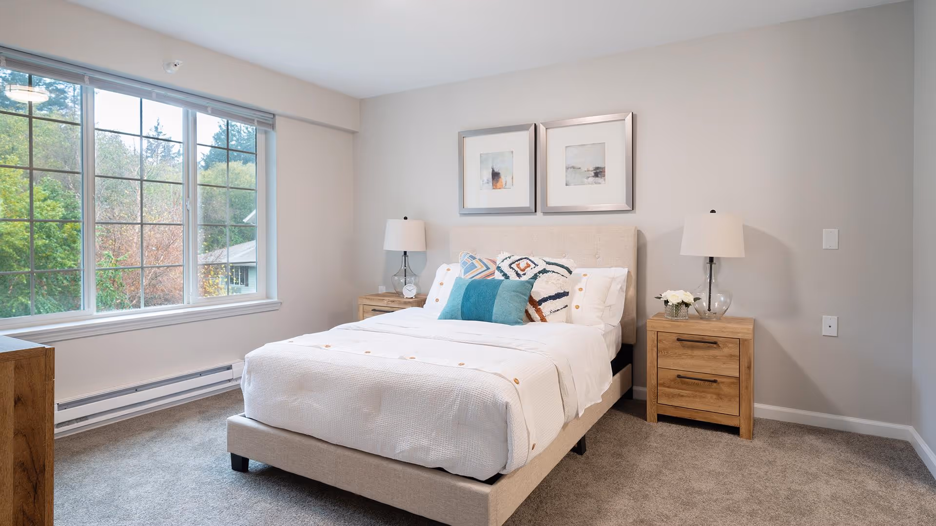 A bright and clean bedroom with a large window showing greenery outside. The room features a neatly made bed with white bedding and decorative pillows, flanked by two wooden nightstands each with a lamp. Two framed abstract artworks hang above the bed.