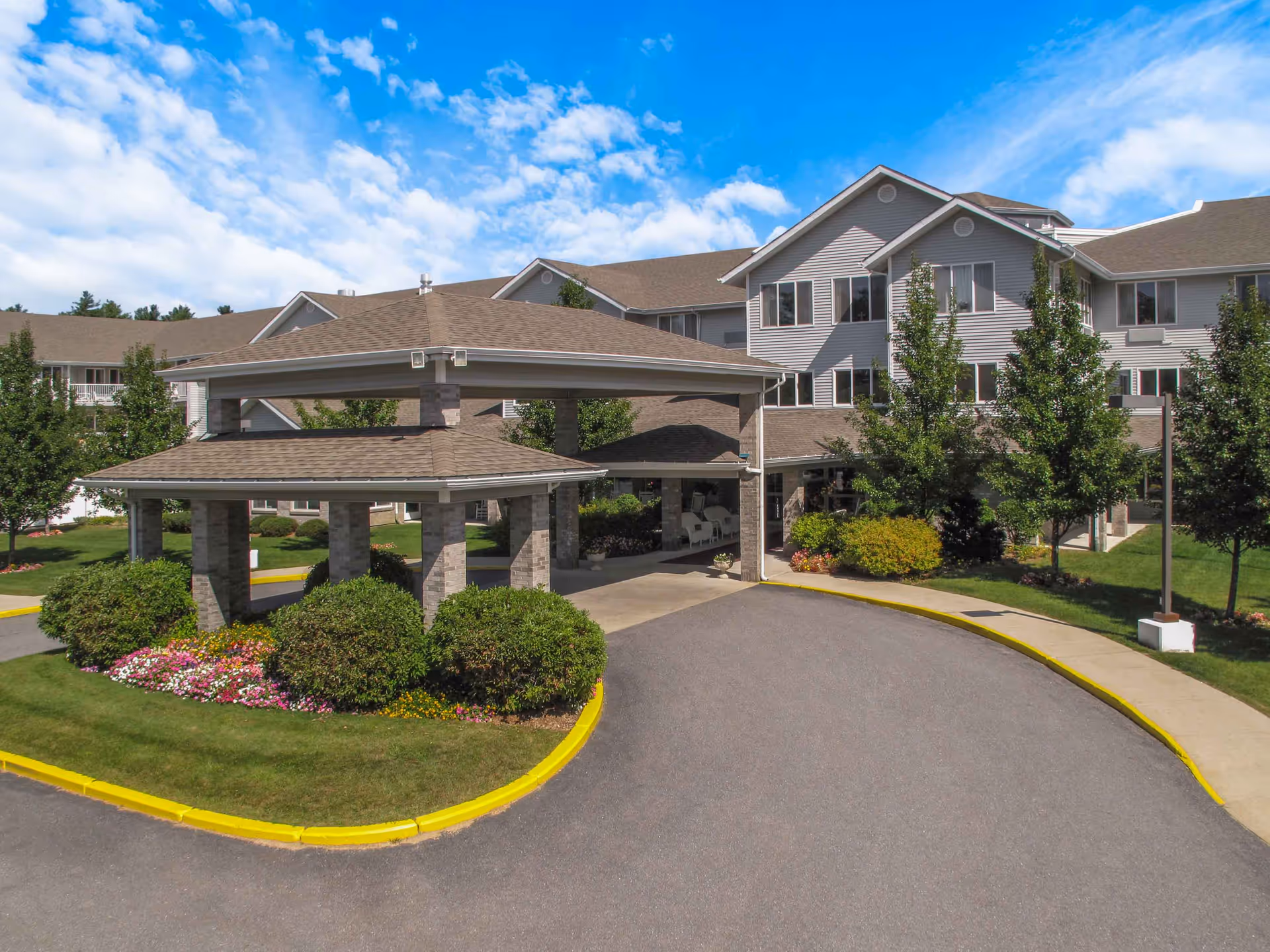 Exterior view of Holiday Woods at Canco senior living facility showing a large covered entrance with stone pillars, surrounded by well-maintained landscaping including bushes and flowers, under a partly cloudy blue sky.
