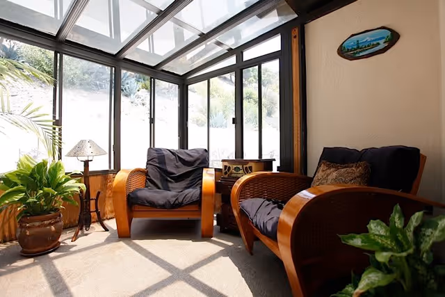A sunlit indoor seating area with two wooden armchairs featuring dark cushions, surrounded by large windows and a glass ceiling. There are two potted plants, a small lamp on a stand, and a small wooden table with a decorative item between the chairs. A small wall decoration hangs on the beige wall.