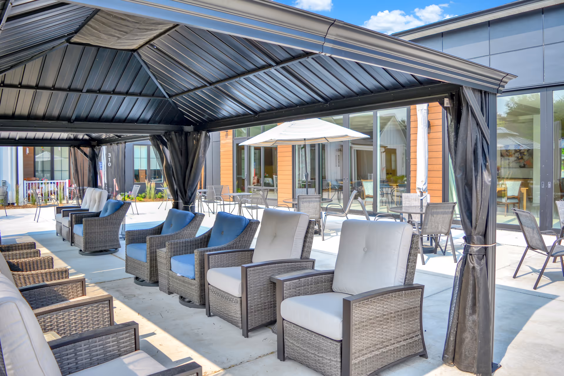 Outdoor seating area at The Woods - A GreatLife Community featuring cushioned wicker chairs arranged under a large metal canopy. In the background, there are additional tables and chairs with umbrellas on a concrete patio adjacent to a building with large glass windows and doors.