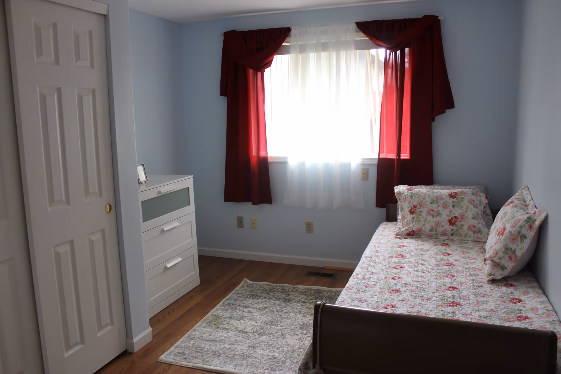 A small bedroom with light blue walls, a single bed with floral bedding and pillows, a white dresser with three drawers, a window with sheer white curtains and red drapes, and a light-colored patterned rug on a wooden floor.