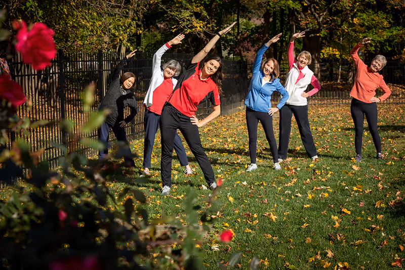A group of six women standing on a grassy area outdoors, performing a side stretch exercise with one arm raised overhead and the other on their hip. They are dressed in casual athletic clothing and appear to be enjoying a sunny day surrounded by trees and fallen leaves.