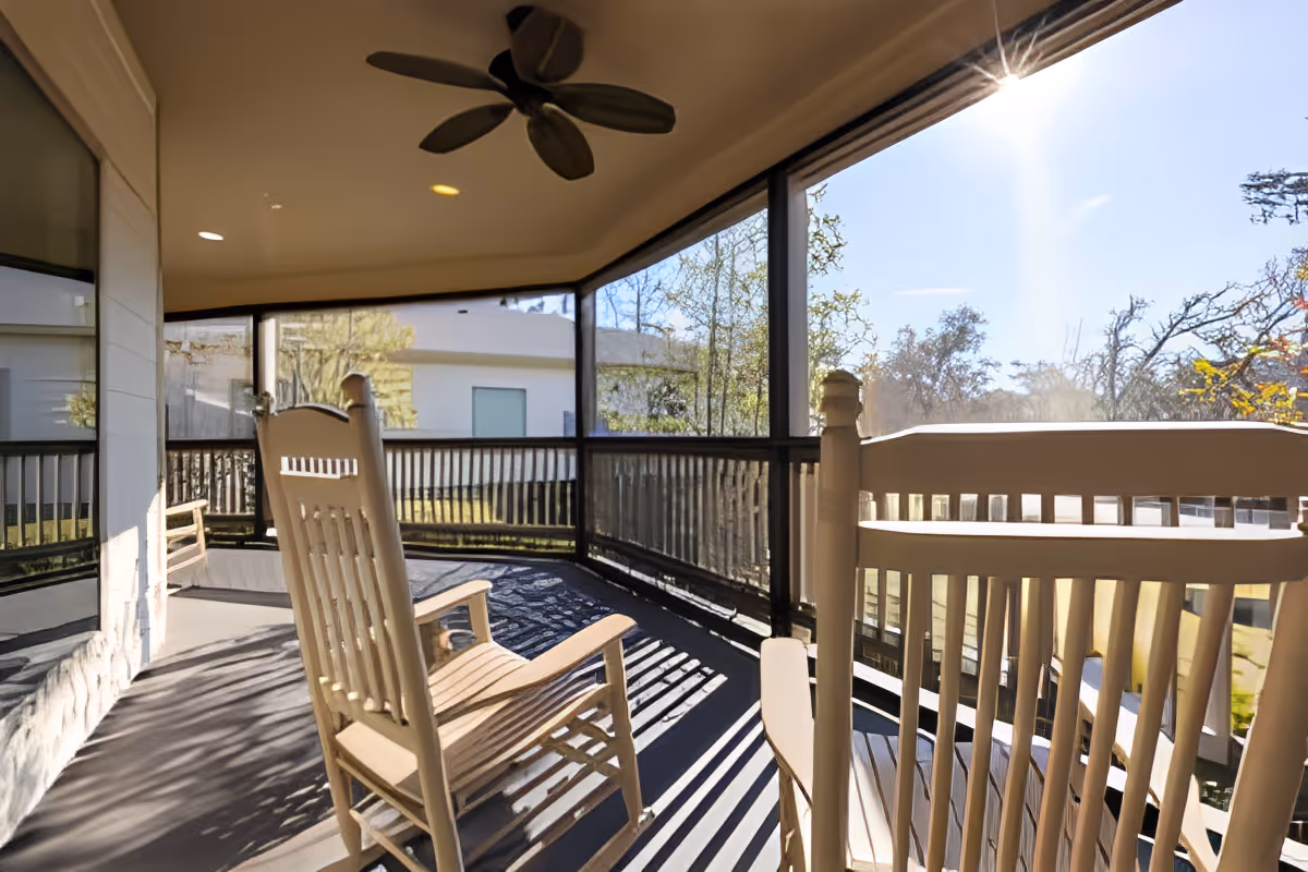Covered screened porch with wooden rocking chairs and a ceiling fan overlooking a sunny yard.