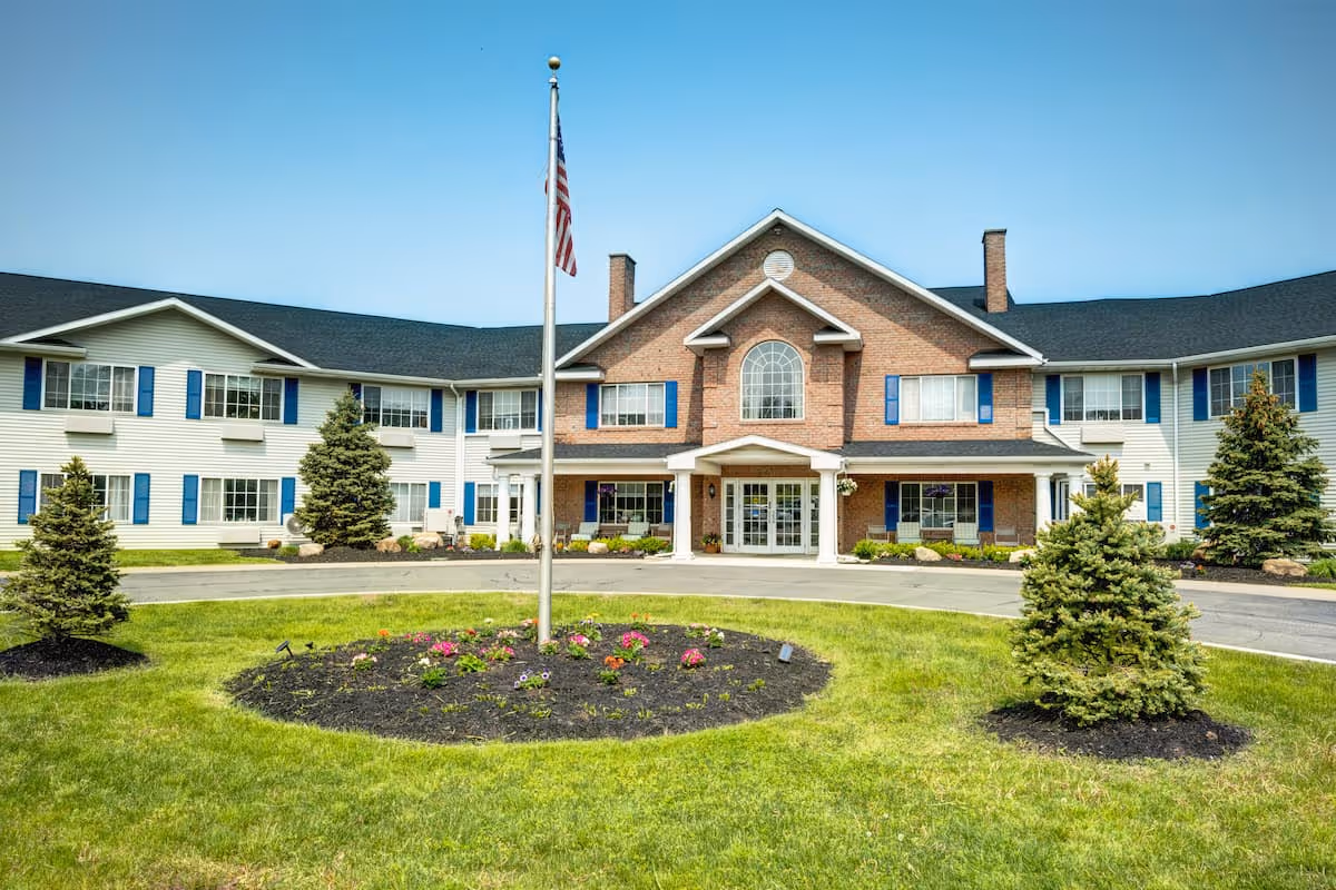 Front exterior view of Evergreen Place, a two-story senior living facility with a brick and white siding facade, blue shutters, a central entrance with a covered porch, an American flag on a flagpole in front, and landscaped greenery including small trees and a flower bed.