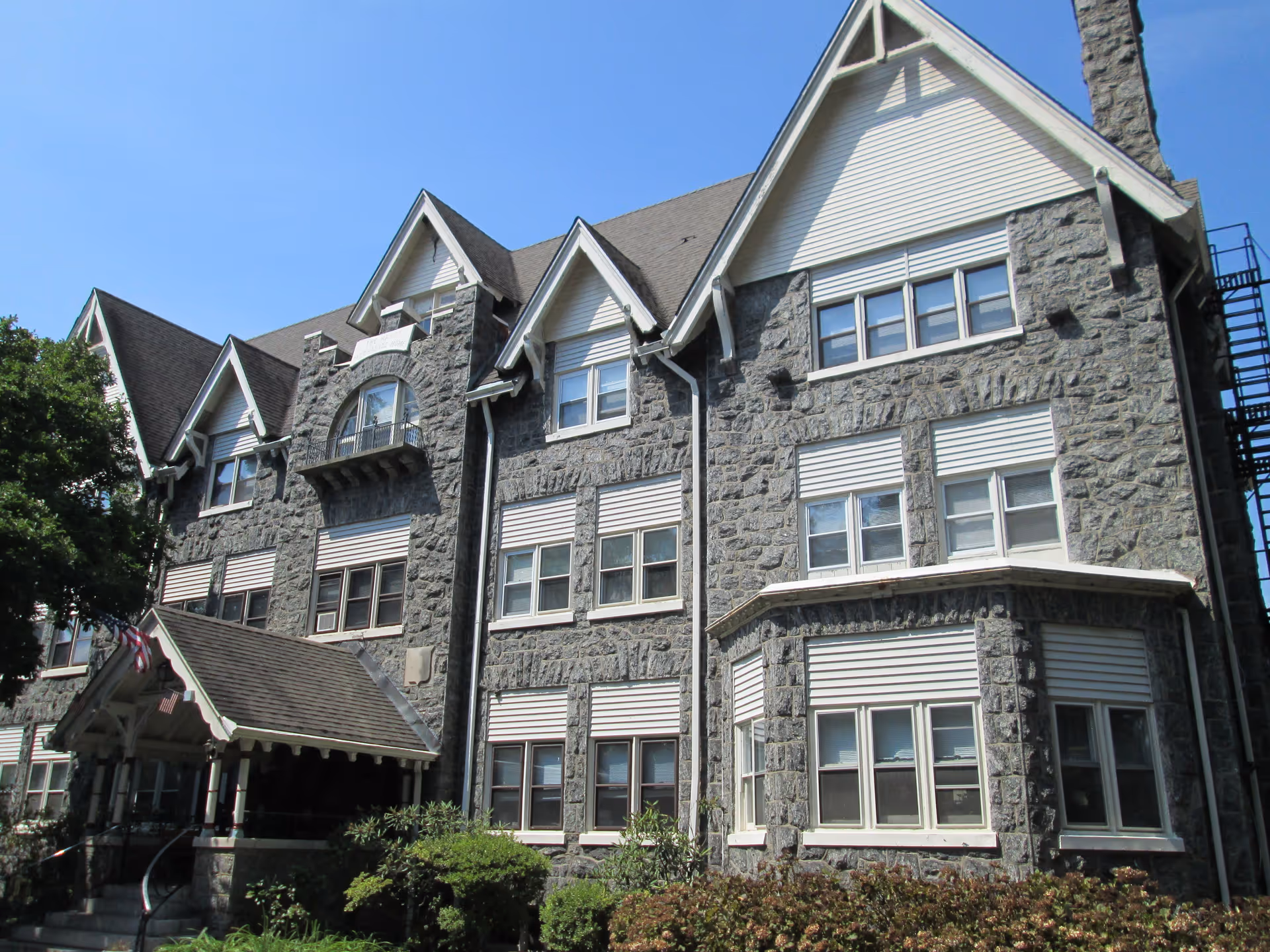 Exterior view of a large stone building with multiple windows and a steeply pitched roof under a clear blue sky. The building has a covered entrance with an American flag and surrounding greenery.