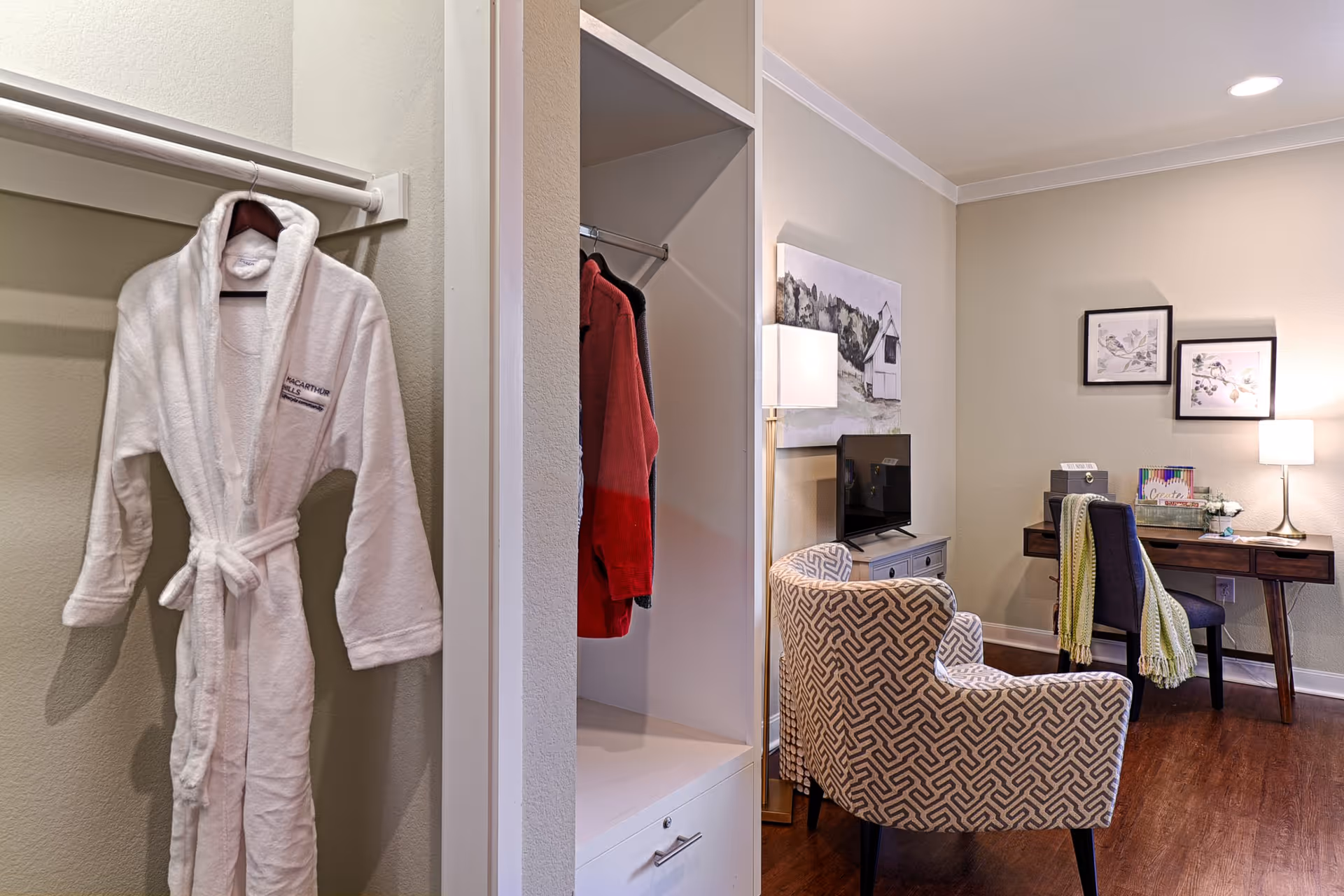 Interior view of a senior living facility room showing a white bathrobe hanging in an open closet on the left, a patterned armchair facing a small TV on a cabinet, and a wooden desk with a chair, lamp, and decorative items on the right. The walls are light beige with framed artwork, and the floor is wooden.