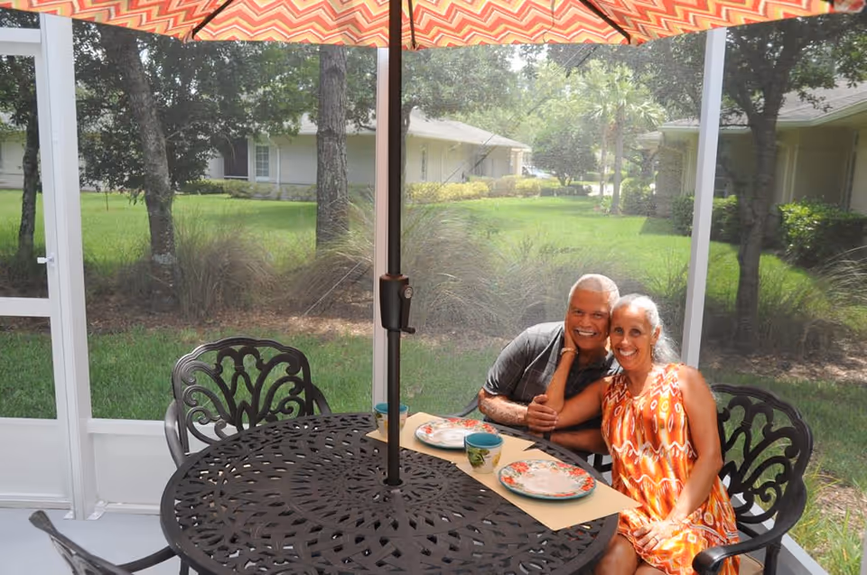 An elderly couple sitting closely together and smiling at a round outdoor metal table with two plates and two cups under a large patterned umbrella. They are in a screened patio area with green grass, trees, and residential buildings visible outside.