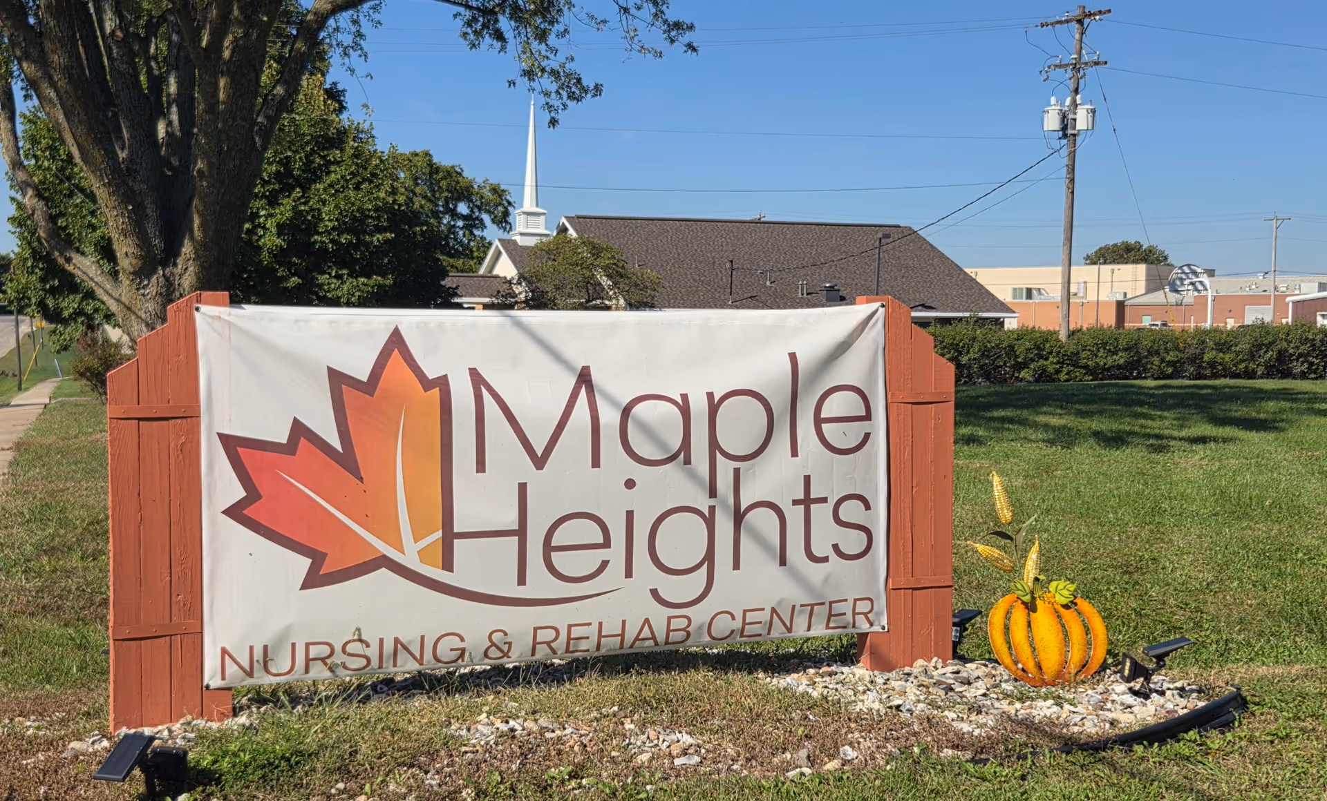 Outdoor sign reading "Maple Heights Nursing & Rehab Center" with a leaf logo and a pumpkin decoration on a lawn in front of the facility.