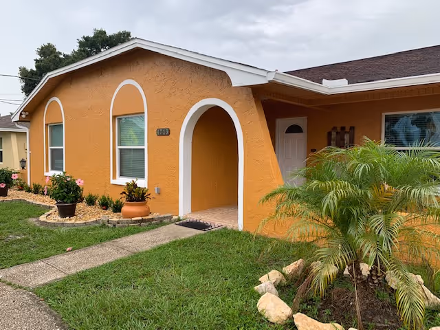 Exterior view of a single-story orange building with white trim, featuring an arched entrance and two windows. There are potted plants and landscaping with green grass and small bushes in front of the building under a cloudy sky.