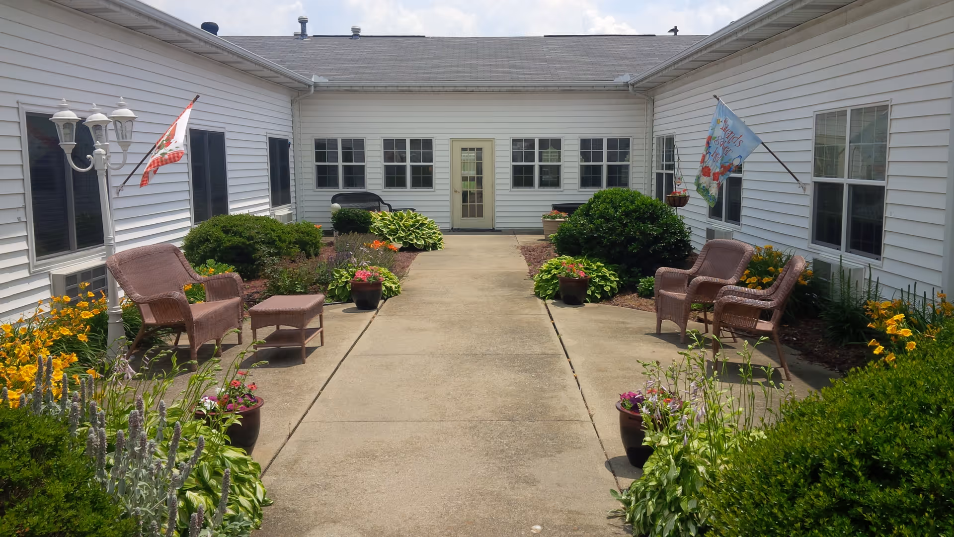 Outdoor courtyard area of a senior living facility with white siding buildings on three sides, a concrete walkway in the center, wicker chairs and ottomans on both sides, potted plants, bushes, flowers, and two flags mounted on the building walls.