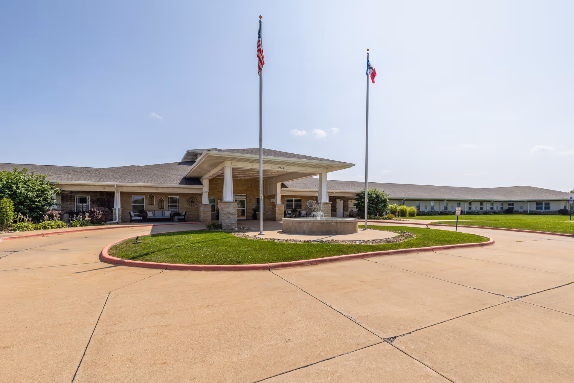 Main entrance of a senior living facility with a covered porte-cochère, circular driveway, central fountain, and two flagpoles.