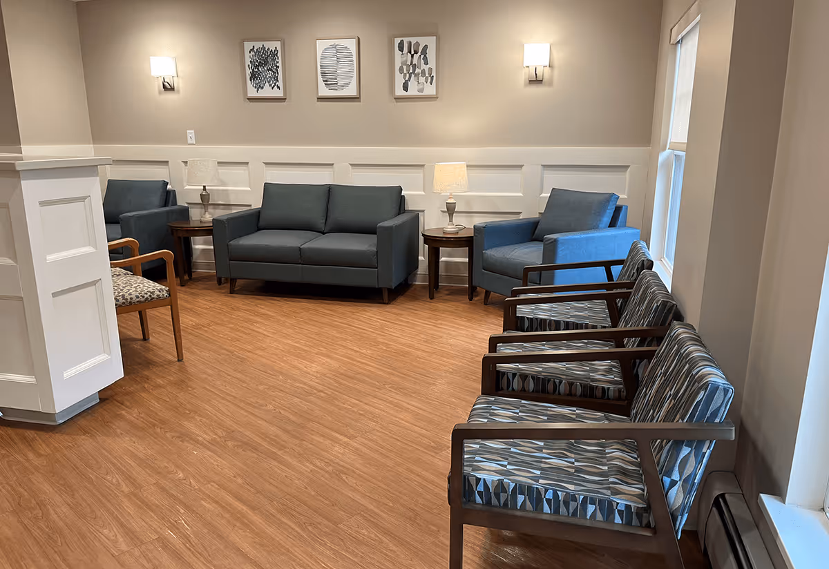 A cozy senior living community lounge area with wooden flooring, two blue armchairs, a blue loveseat, three patterned chairs, two side tables with lamps, and three abstract framed artworks on the wall. The room is softly lit with wall sconces and natural light from windows.
