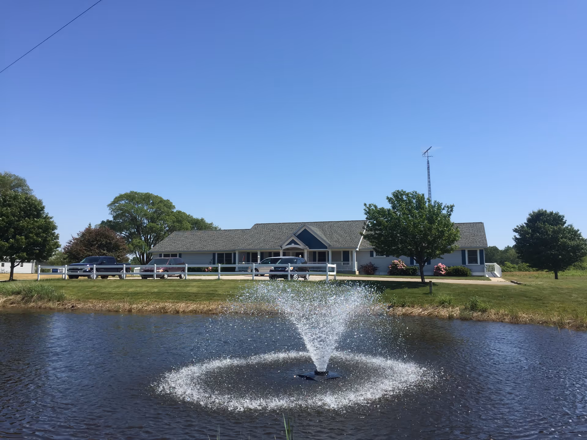 A single-story building with a gray roof and white exterior, surrounded by green grass and trees under a clear blue sky. In the foreground, there is a pond with a water fountain spraying water upwards. Several cars are parked in front of the building behind a white fence.