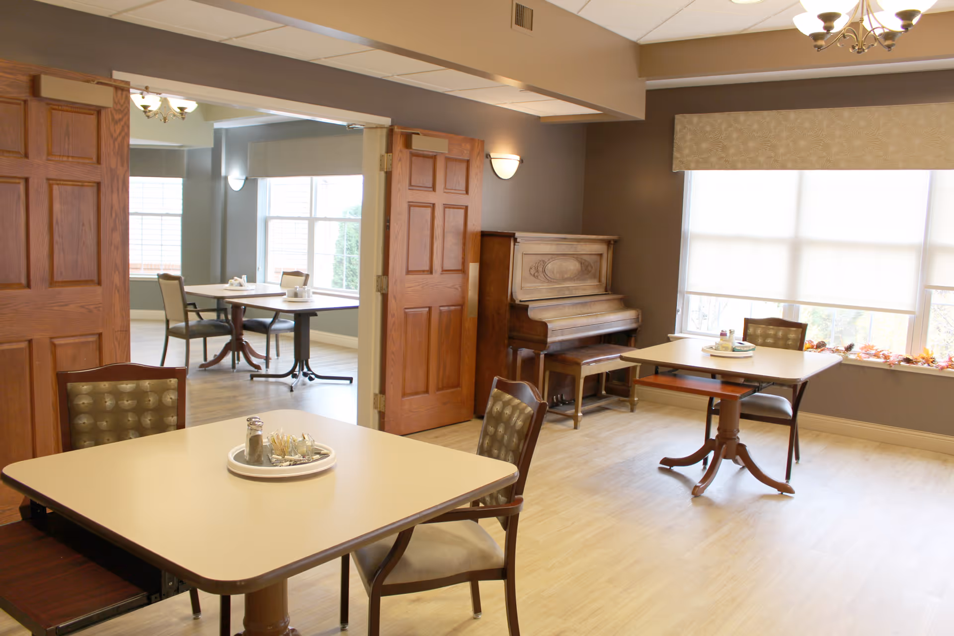 A bright and clean dining area in a senior living facility with several tables and chairs. The room features large windows with blinds, wooden double doors, and a piano against one wall. The tables have small condiment trays on them.