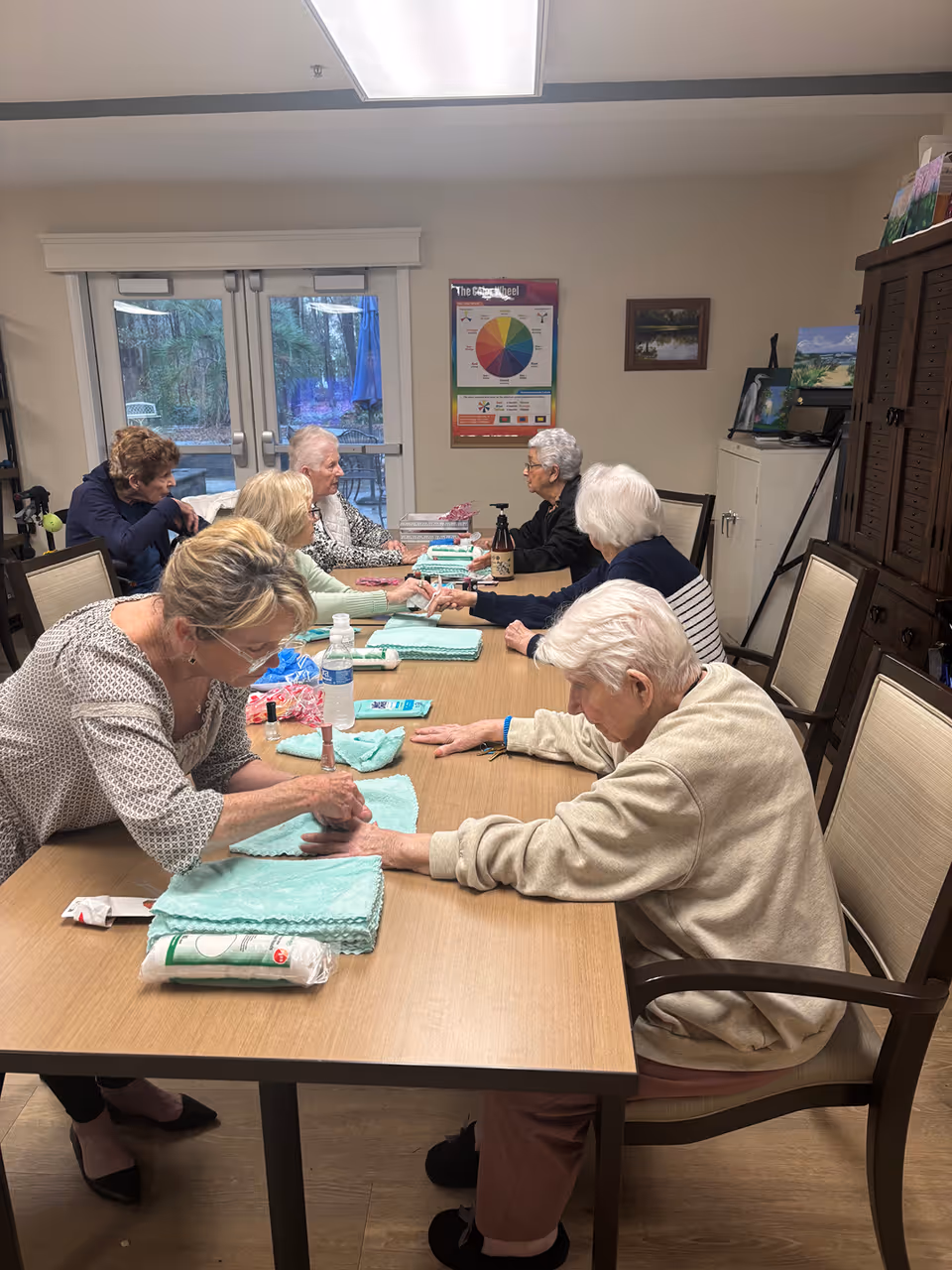 A group of elderly women sitting around a rectangular table in a well-lit room, engaging in a manicure activity. One woman is applying nail polish to another's nails while others wait or chat. The room has a large window with a view of trees outside, a colorful chart on the wall, and various furniture including chairs and cabinets.