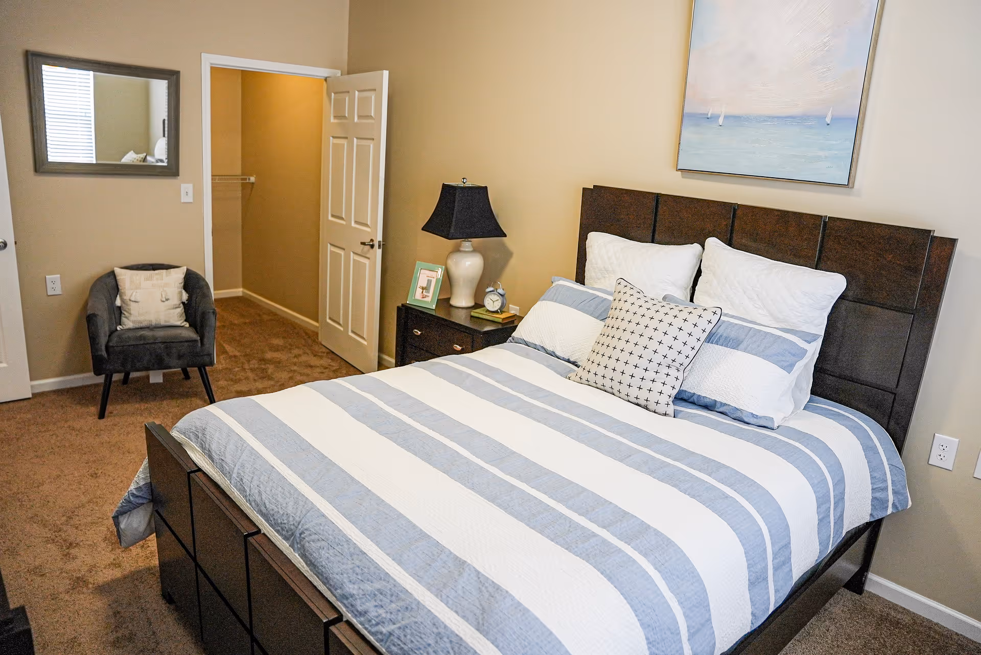 Well-lit bedroom with a striped bed and dark wood headboard, nightstand with a lamp, an armchair, and an open closet door.