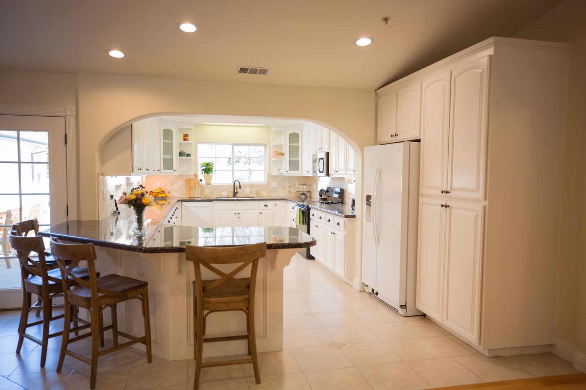 Bright kitchen with white cabinets, a large window above the sink, a granite countertop island with three wooden chairs, and modern appliances including a refrigerator, stove, and microwave.