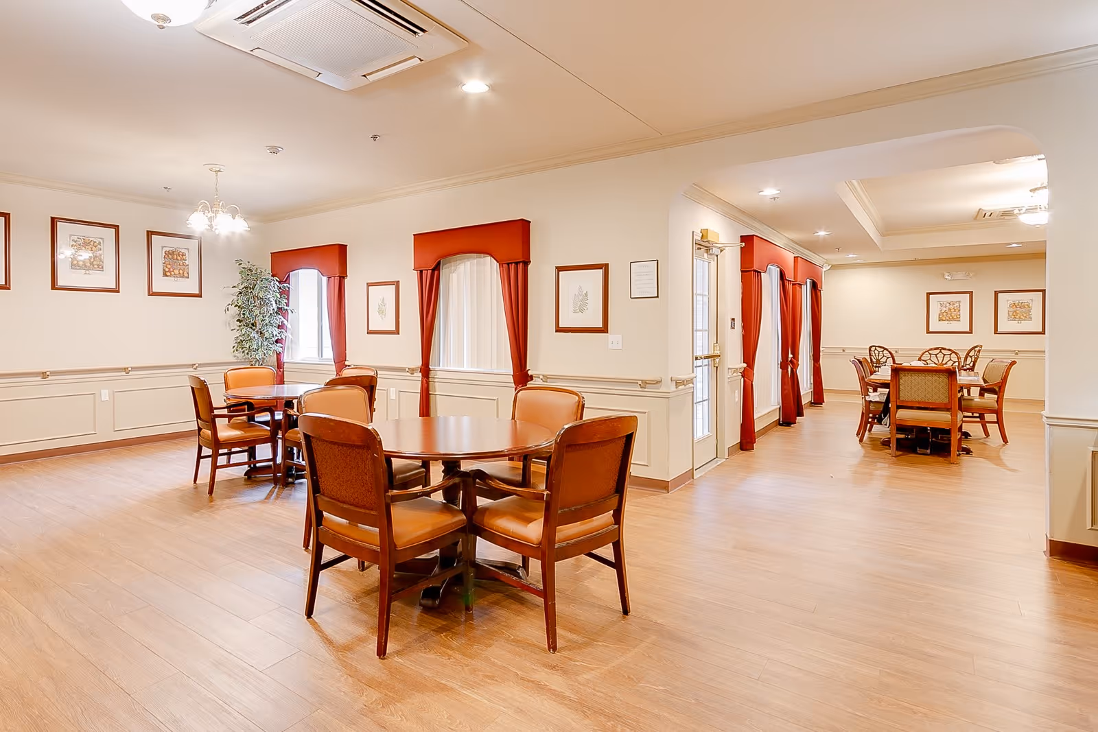 A bright and spacious dining room in a senior living facility with wooden floors, round tables, and cushioned chairs. The walls are decorated with framed artwork and windows are adorned with red curtains. The room is well-lit with ceiling lights and has a clean, welcoming atmosphere.